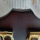 Detail hlavy tmavě hnědé dřevěné kytary s kovovými zlatými ladičkami. Na horním okraji hlavy je viditelné odloupaní laku a poškození povrchu. Na fotc…