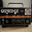 Černý kytarový zesilovač s kovovou rukojetí a oranžovým pruhem na předním panelu. Na horní části je bílý nápis ORANGE a vedle něj text MICRO DARK, v…