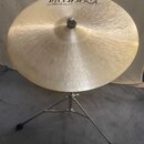 Zlatý kovový činel umístěný na chromovaném stativu. Na povrchu je nápis „istanbul Mehmet Handmade Cymbals from Turkey“. Povrch činelu vykazuje jemné…