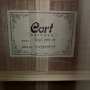 Fotografie zachycuje štítek nalepený na dřevěném povrchu vnitřku nástroje. Štítek nese logo Cort GUITARS, označení MODEL: GA5F FMH OP a SERIAL NO: CA…