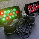 Dvě černé LED par lampy s obdélníkovým tělem na cylindrických základnách. Levá lampa svítí kombinací zeleného a červeného světla, pravá lampa svítí č…