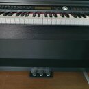 Černý digitální piano s klávesnicí a třemi pedály. Horní panel obsahuje ovládací tlačítka, displej a kulatý ovladač. Na přístroji nejsou viditelné žá…