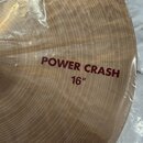Detailní pohled na mosazný činel s viditelným rýhováním povrchu. Na čineli je červeným písmem nápis POWER CRASH 16". Nástroj je zabalen v průhledné p…
