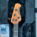 Hlava basové kytary z přírodního dřeva v černém polstrovaném kufru. Na hlavě je černý nápis „ERNIE BALL MUSICMAN StingRay“. Jsou zde čtyři černé ladi…