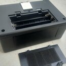 Černý plastový box s otevřeným přihrádkou pro baterie, ve které jsou vidět kovové pružiny. Vedle boxu leží oddělený černý plastový víčko s vnitřní pě…