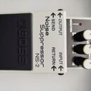 Bílý efekt pedál s černým vypínačem. Na šasi je uveden nápis Noise Suppressor NS-2 a označení vstupů a výstupů OUTPUT, SEND, RETURN, INPUT. Na černém…