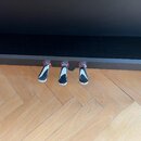 Detailní pohled na tři kovové pedály pianina v chromovém provedení. Pedály jsou umístěny v dolní části černého nástroje s matným povrchem. Nástroj st…