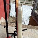 Fotografie zachycuje elektrickou kytaru v profile, umístěnou na černém stojanu s červeným základnám. Tělo kytary je v barevné variantě stříbrný metal…