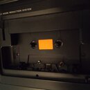 Fotografie zachycuje otevřený přehrávač kazet v černé barvě. Na pravé straně je bílý nápis DBX NOISE REDUCTION SYSTEM. V prostoru pro kazetu je vidět…
