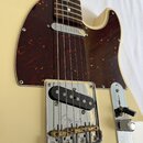 Detail elektrické kytary krémové barvy s hnědým želvovinovým pickguardem. Na chromovaném krytu snímače je vyryto logo Fender. Nástroj má jeden černý…