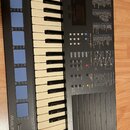 Šedočerný digitální klavír s bílými a černými klávesami. Na levé straně jsou modré gumové podložky a nápisy PERCUSSION SET A, CHORD START a logo Musi…