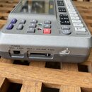 Stříbrno-šedý elektronický hudební přístroj s displejem a řadou tlačítek. Na boku jsou nápisy POWER ON • OFF a MAX VOLUME MIN s vypínačem a posuvníke…