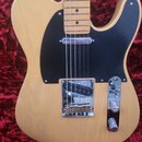 Elektrická kytara s přírodním dřevěným povrchem těla a světle hnědým hmatníkem. Nástroj má černý pickguard, jeden ovládací knoflík a chromovaný most…