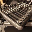 Stříbrno-černý mixážní pult s řadou posuvníků, knoflíků a tlačítek. Na horním panelu je vidět bílý nápis X-TOUCH. Zařízení je umístěno na hliníkovém…