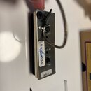 Fotografie zachycuje spodní stranu elektrického snímače s kovovým krytem stříbrné barvy. Na spodní části je nalepený bílý štítek s nápisem NECK a ved…