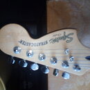 Fotografie zachycuje hlavu elektrické kytary z přírodního světlého dřeva. Na hlavě je vidět nápis Squier a STRATOCASTER®. Jsou zde vidět šest chromov…