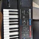 Černý digitální klávesový nástroj s bílými a černými klávesami. Na ovládacím panelu jsou nápisy D-BEAM, VOLUME, MODE/MIC a KEYBOARD. Jsou zde viditel…