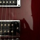 Detail korpusu elektrické kytary v tmavě červené barvě s lesklým povrchem. Jsou vidět dva humbuckery s chromovanými rámečky a černými snímači s vidit…