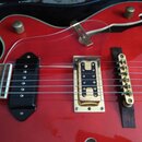 Detail červené polodubové elektrické kytary s průhledným pickguardem. Nástroj má dva snímače typu humbucker, z nichž jeden je černý a druhý v kovovém…