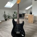 Černá elektrická kytara typu Stratocaster s černým pickguardem a hnedým hmatníkem. Na hlavě je viditelné logo „Fender Stratocaster“. Nástroj je umíst…