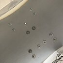 Detailní pohled na šedý, hladký povrch s rozmístěnými kovovými šrouby a vruty. V pravém dolním rohu je vidět část bílého povrchu s černým nápisem „RE…