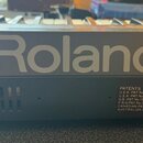Detailní pohled na část klávesového nástroje s velkým šedým nápisem Roland. Pod ním je černý panel s ventilací a štítkem PATENTS, který obsahuje sezn…