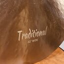 Detailní záběr na mosazný činel s viditelnou strukturou kování a drobnými nečistotami na povrchu. Uprostřed je bílý nápis „Traditional“ a pod ním tex…