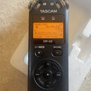 Černý digitální záznamový zařízení s nápisem TASCAM a označením modelu DR-05. Displej je zapnutý s oranžovým podsvícením a zobrazeným textem 00H00M00…