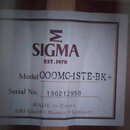 Fotografie zachycuje štítek uvnitř akustické kytary. Štítek obsahuje logo s řeckým písmenem sigma a nápisy SIGMA, EST.1970, Model 000MC-1STE-BK+, Ser…