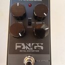 Černý efekt pedál s kovovým tělem a čtyřmi ovládacími knoflíky. Na povrchu jsou nápisy „tc electronic“, „FANGS METAL DISTORTION“ a „TRUE BYPASS“. Mez…