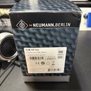 Fotografie znázorňuje černou papírovou krabici s logem a nápisem NEUMANN.BERLIN. Na krabici je bílá nálepka s textem TLM 49 Set, Kondensator-Mikropho…
