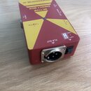 Červená aktivní DI box s žlutými trojúhelníkovými prvky na horní straně. Na zařízení jsou nápisy ACTIVE DIRECT BOX, PROFESSIONAL DI, LINE IN, PARALLE…