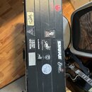 Černý tvrdý kufýr s několika nálepkami. Jsou na něm viditelné loga a nápisy: ROGER, SHURE, Elixir STRINGS, tc electronic, Eventide Devices a nápis IF…