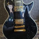Černá elektrická kytara s lesklým povrchem a bílým lemováním. Nástroj má dva zlaté humbuckery, zlatý kobylník a čtyři černé oválné knofíky. Na povrch…