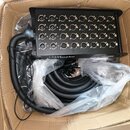 Černý vícežilový kabel s konektorovým boxem umístěný v kartonové krabici. Box obsahuje 32 female XLR konektorů uspořádaných do čtyř řad po osm. Na sp…