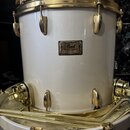 Bílý buben s pozlaceným kováním. Na korpusu je kovový štítek s nápisy „Pearl“, „Masters Custom“, „Custom Series“ a „MADE IN JAPAN“. Povrch nástroje i…