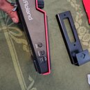 Černý elektronický přístroj s růžovými hranami. Na horní části je bílý nápis Roland. Na boku jsou umístěna dvě kulatá tlačítka s popisky SENS a THRES…