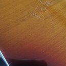 Detailní záběr na lakovaný dřevěný povrch hudebního nástroje v červeno-žlutém přechodu (sunburst). V horní části jsou vidět výrazné povrchové škrában…