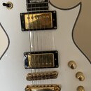 Detail bílé elektrické kytary s gold hardwarem. Nástroj má dva humbuckery se zlatými kryty na černých rámečcích, zlatý kobylník a tři zlaté oválné kn…