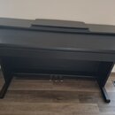 Černý digitální piano v konzolové podobě s uzavřenou klaviaturowou víkem. Nástroj stojí na dvou širokých nohách a v dolní části jsou vidět tři pedály…