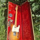 Elektrická kytara s tvarem typu Telecaster v hnědém sunburst provedení a bílým pickguardem. Nástroj je umístěn v otevřeném kufru s červenou plyšovou…