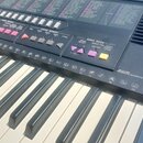 Detailní pohled na ovládací panel a klávesy elektronického klavíru v černé barvě. Na panelu jsou vidět nápisy VOICE EFFECT, CHORD MEMORY a SONG BOOK,…