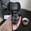 Černý mikrofon značky ZOOM s nasazenou větrolamicí. Na spodní části jsou dvě vstupní zdařila, označená číslicemi 1 a 2, svítící zelená kontrolka CHG…