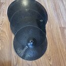 Tři černé perforované kovové disky různých velikostí, položené přes sebe na dřevěné podlaze. Disky mají povrch s hustou mřížkou malých otvorů a v cen…