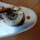 Detailní pohled na elektronickou součástku, pravděpodobně potenciometr, s kovovým tělem a připojeným hnědým kabelem. Na kovovém povrchu je vyražen ná…