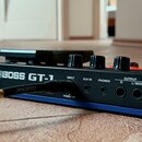 Detailní pohled na zadní panel modrého kytarového procesoru s nápisem BOSS GT-1. Jsou vidět konektory s popisky INPUT, AUX IN, PHONES, OUTPUT R a L/M…