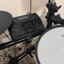 Černý ovládací modul elektronických bicích s řadou tlačítek a posuvníků. Na horní části je nápis Millennium Percussion Drum Module. V dolní části je…
