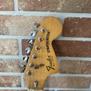 Fotografie zachycuje hlavu elektrické kytary z přírodního dřeva. Na povrchu je černý nápis Fender STRATOCASTER a menší text MADE IN USA. Pod ním je u…