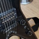 Detailní pohled na černou elektrickou kytaru s nápisovými knoflíky. Na těle je viditelný humbucker s kovovými póly a most s šesti strunami. Povrch ná…