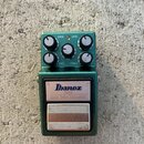 Zelený efekt pedál s kovovým šasi. Na horní části jsou čtyři bílé ovládací knoflíky s popisky DRIVE, LEVEL, BASS a TREBLE a jeden centrální knoflík s…